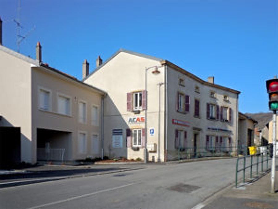 Un grand bâtiment crème à volets bordeaux donne sur une rue de village, avec un petit panneau d'enseigne sur la façade et un feu tricolore au premier plan à droite.