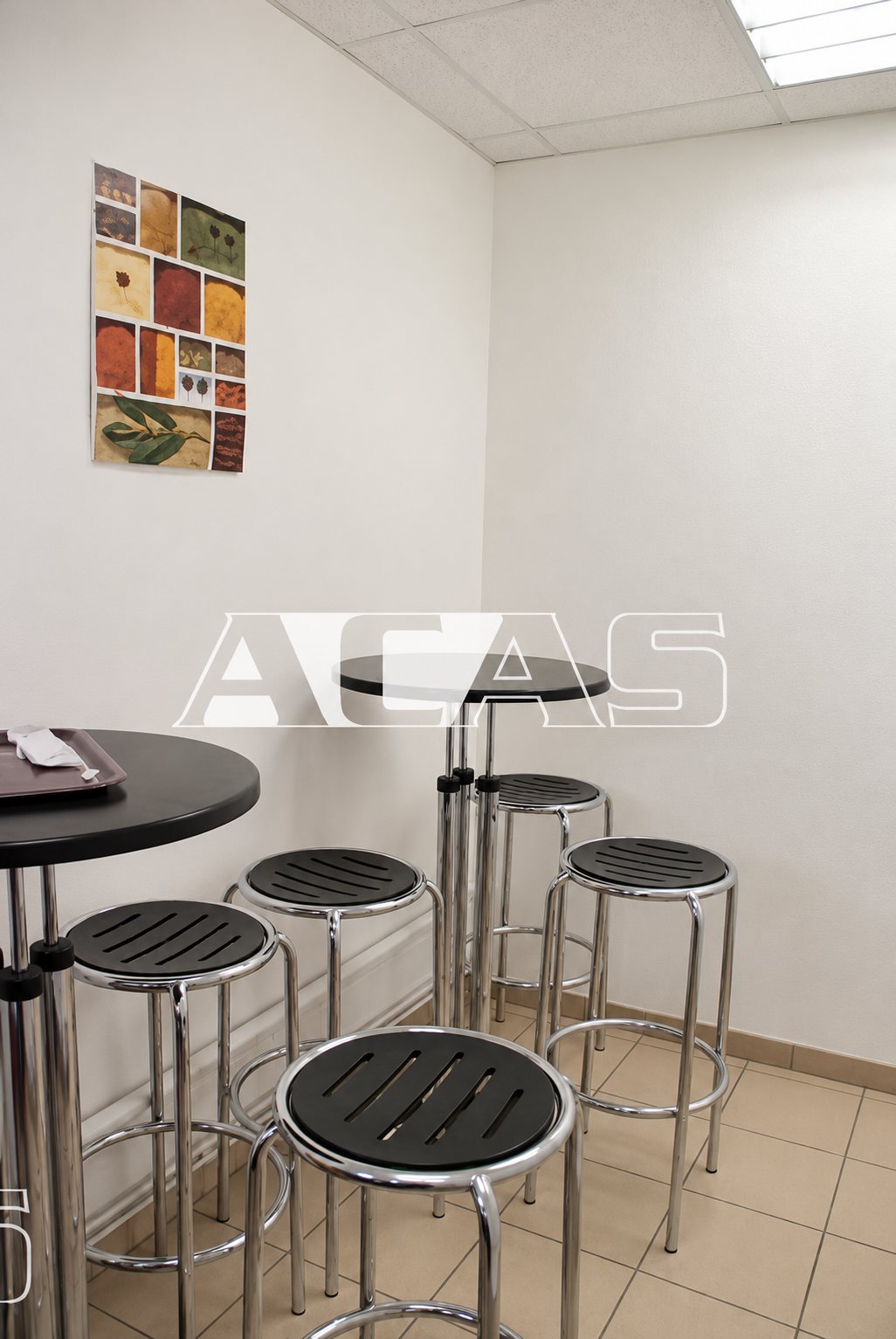 Salle de pause aux murs blancs équipée de tables hautes noires et de tabourets en métal à assise noire, avec un tableau coloré au mur.