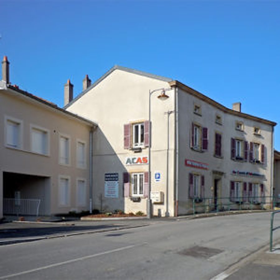 Un bâtiment d'angle beige à volets bordeaux, portant l'enseigne ACAS, donne sur une rue sous un ciel bleu.