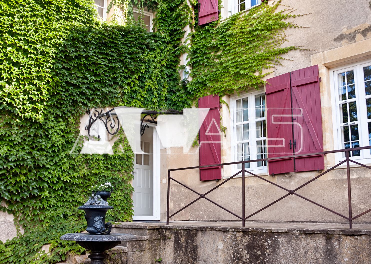 Façade d'une maison ancienne en partie couverte de lierre, avec des volets rouges, une terrasse à balustrade et une petite fontaine au premier plan.