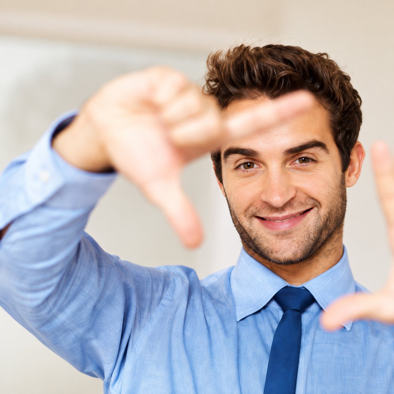 Un homme souriant en chemise bleue et cravate forme un cadre avec ses doigts devant son visage.