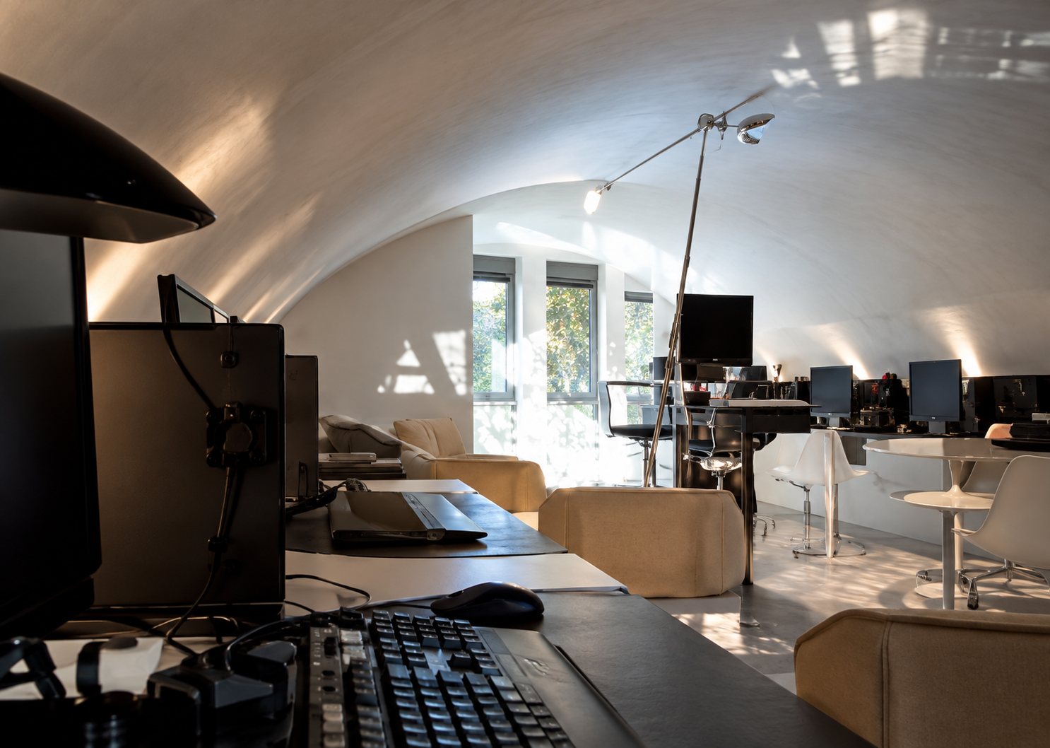 Vue d'un intérieur spacieux à plafond voûté blanc, avec bureaux, chaises et une grande baie lumineuse au fond.