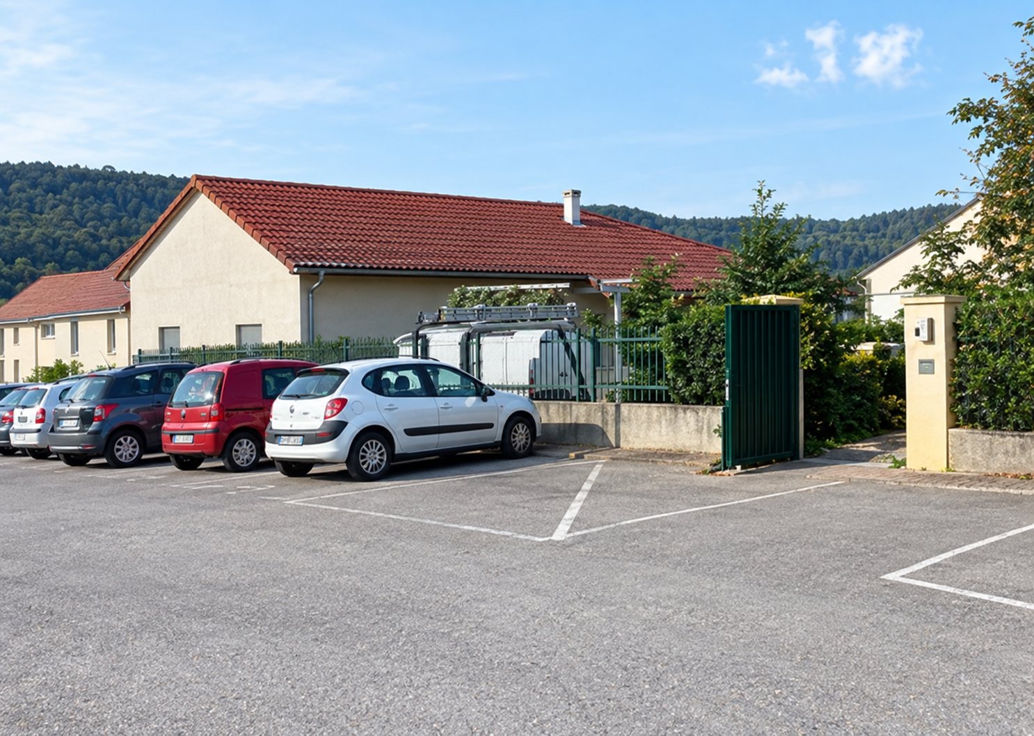 Un parking extérieur où plusieurs voitures sont garées devant un bâtiment bas à toit rouge, avec des collines boisées en arrière-plan.
