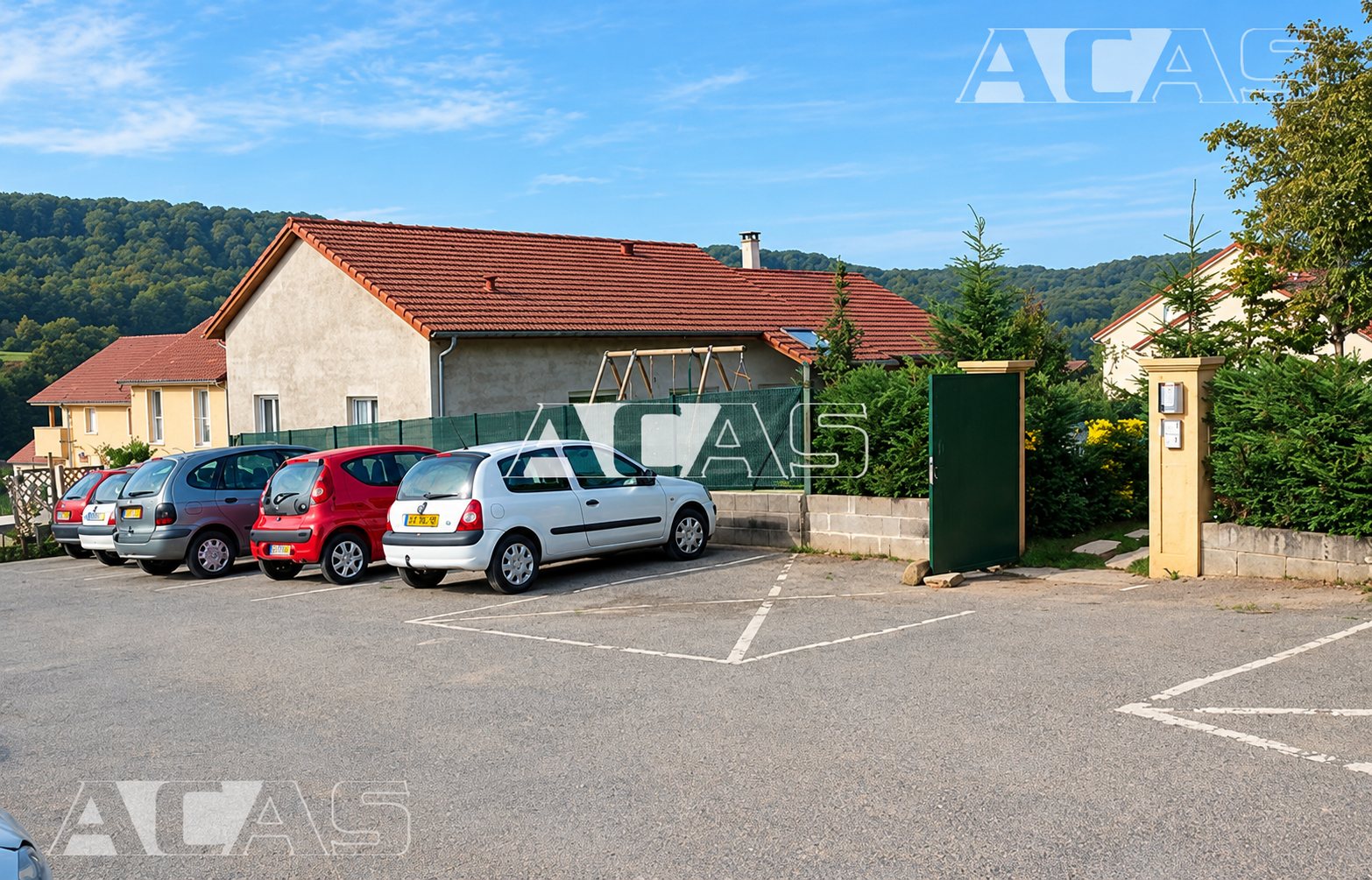 Des voitures sont garées sur un parking goudronné devant une maison en cours de construction à toit de tuiles rouges, avec des collines boisées en arrière-plan.