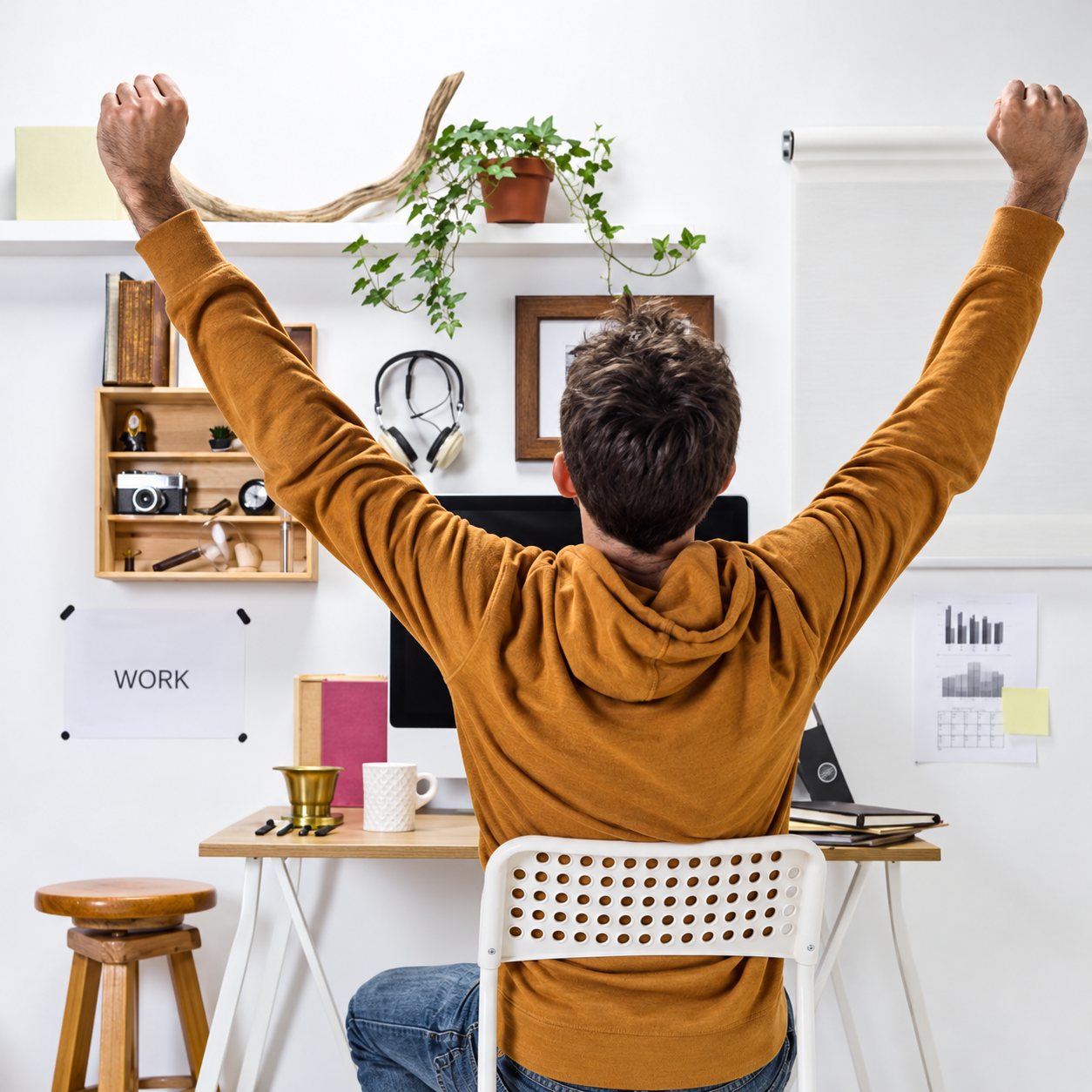Un homme en pull orange, vu de dos, lève les bras de satisfaction devant son bureau blanc, entouré d'étagères, de plantes et d'affiches.