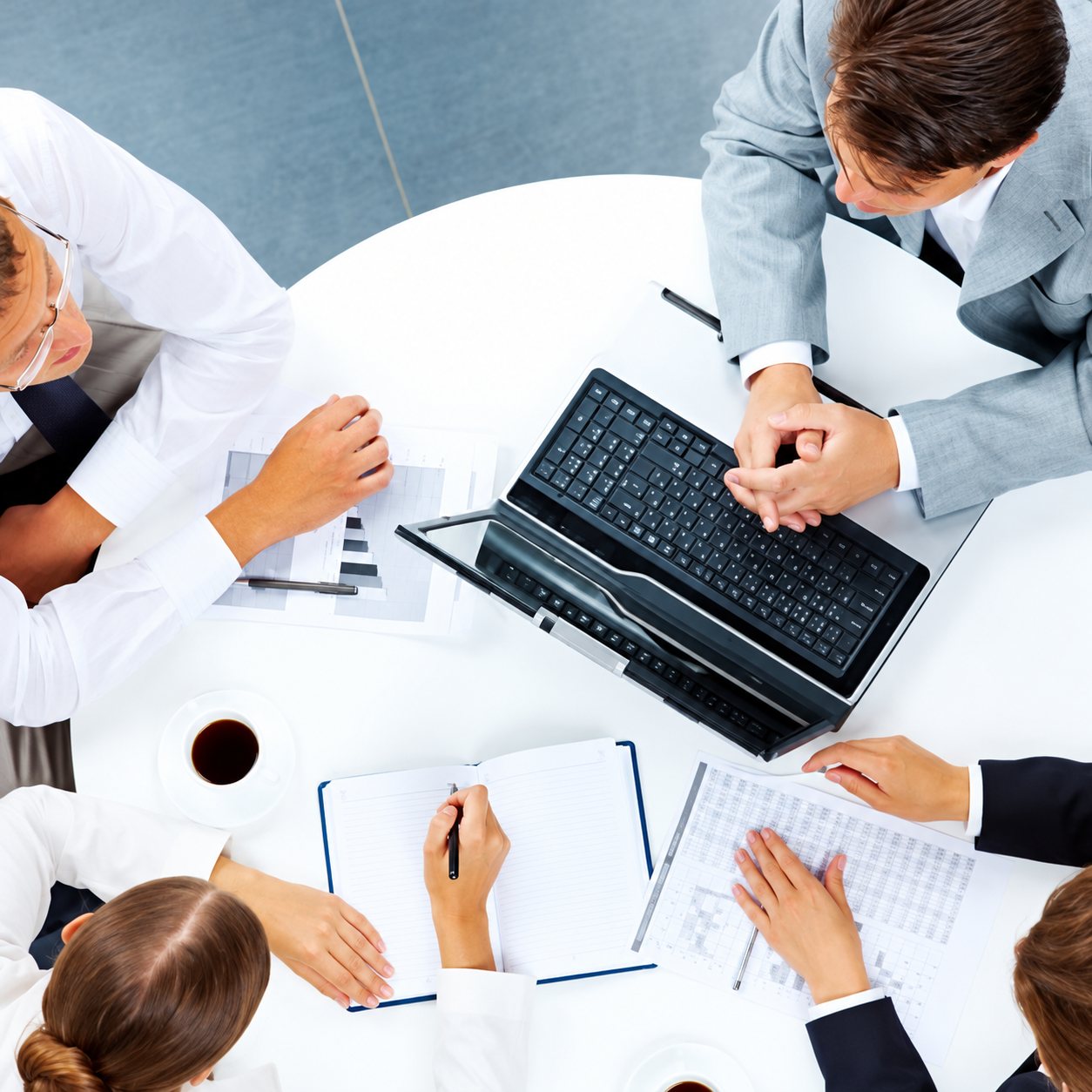 Vue du dessus de plusieurs personnes en tenue professionnelle réunies autour d'une table blanche avec un ordinateur portable, des carnets et une tasse de café.