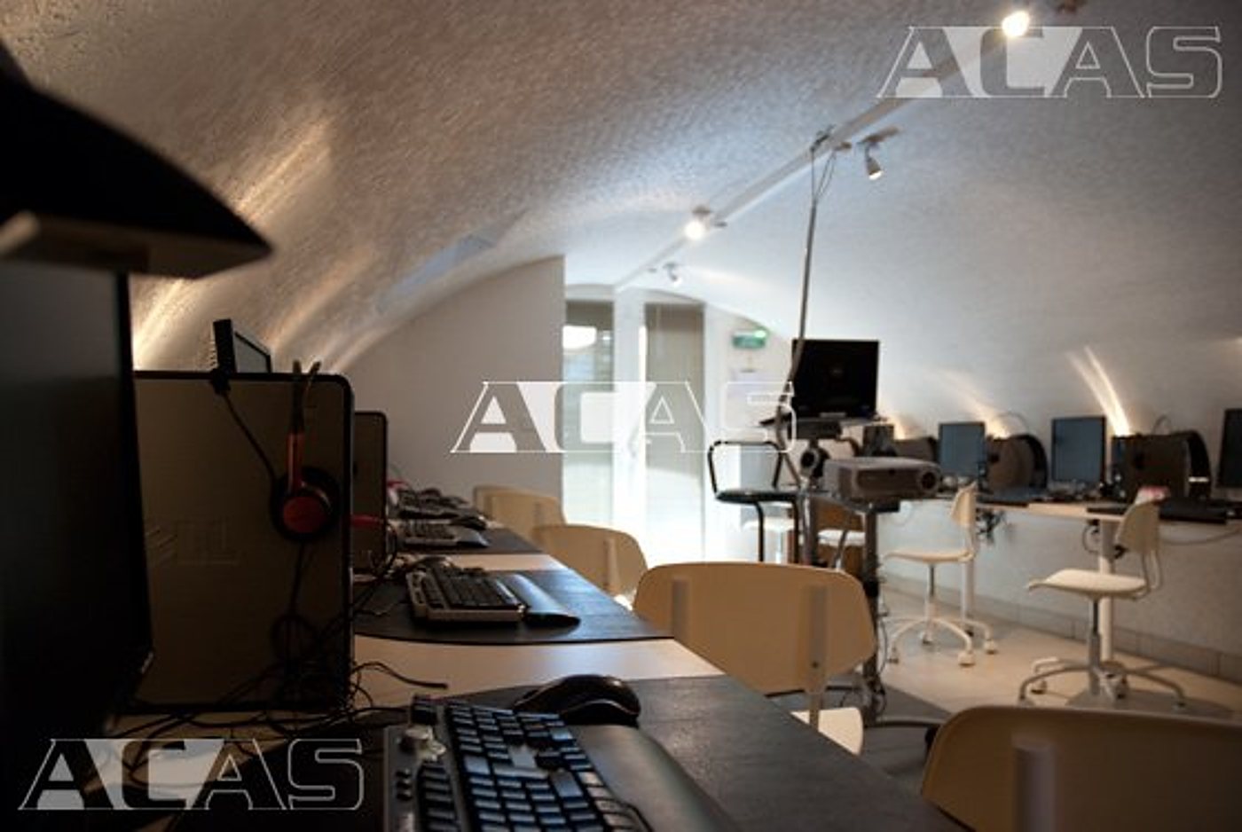 Une salle informatique aménagée sous une voûte claire, avec des claviers, des écrans, un projecteur et des chaises en bois alignés le long des tables.