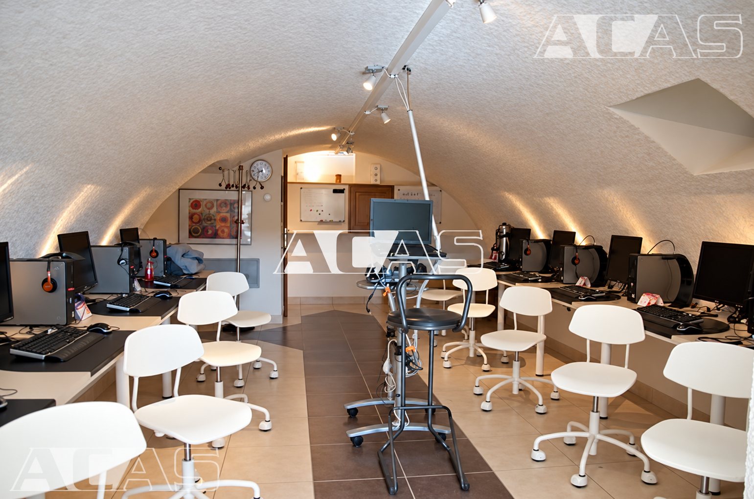 Salle voûtée en pierre claire équipée de postes informatiques alignés le long des murs et de chaises blanches à roulettes.