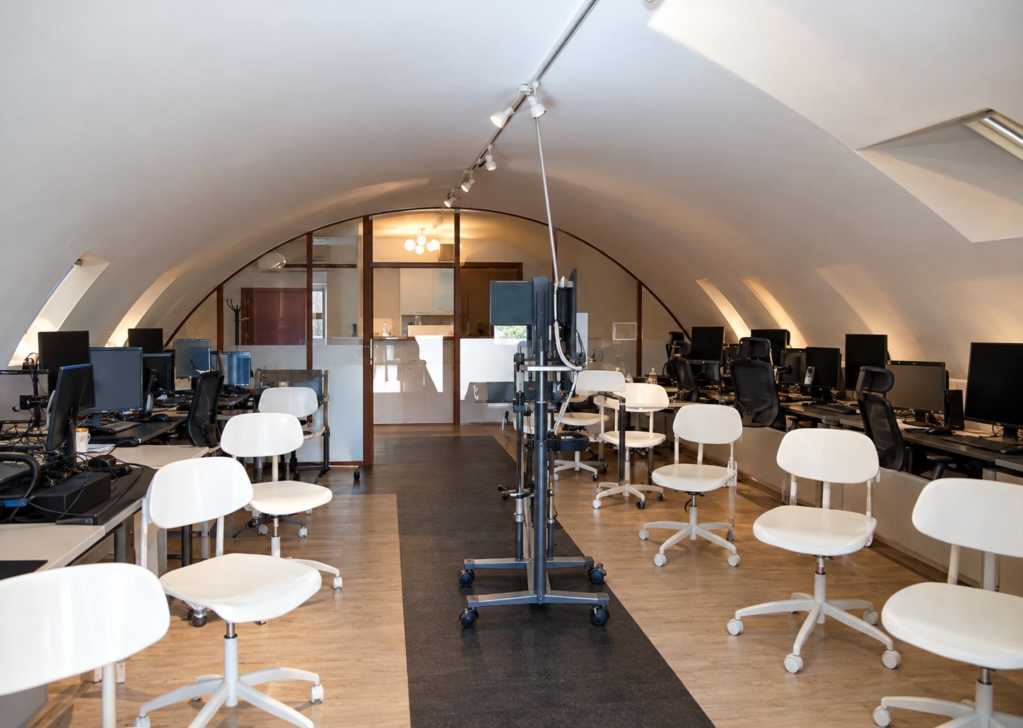 Intérieur d'une salle voûtée aux murs clairs, équipée de chaises blanches et de fauteuils noirs devant des postes de travail.